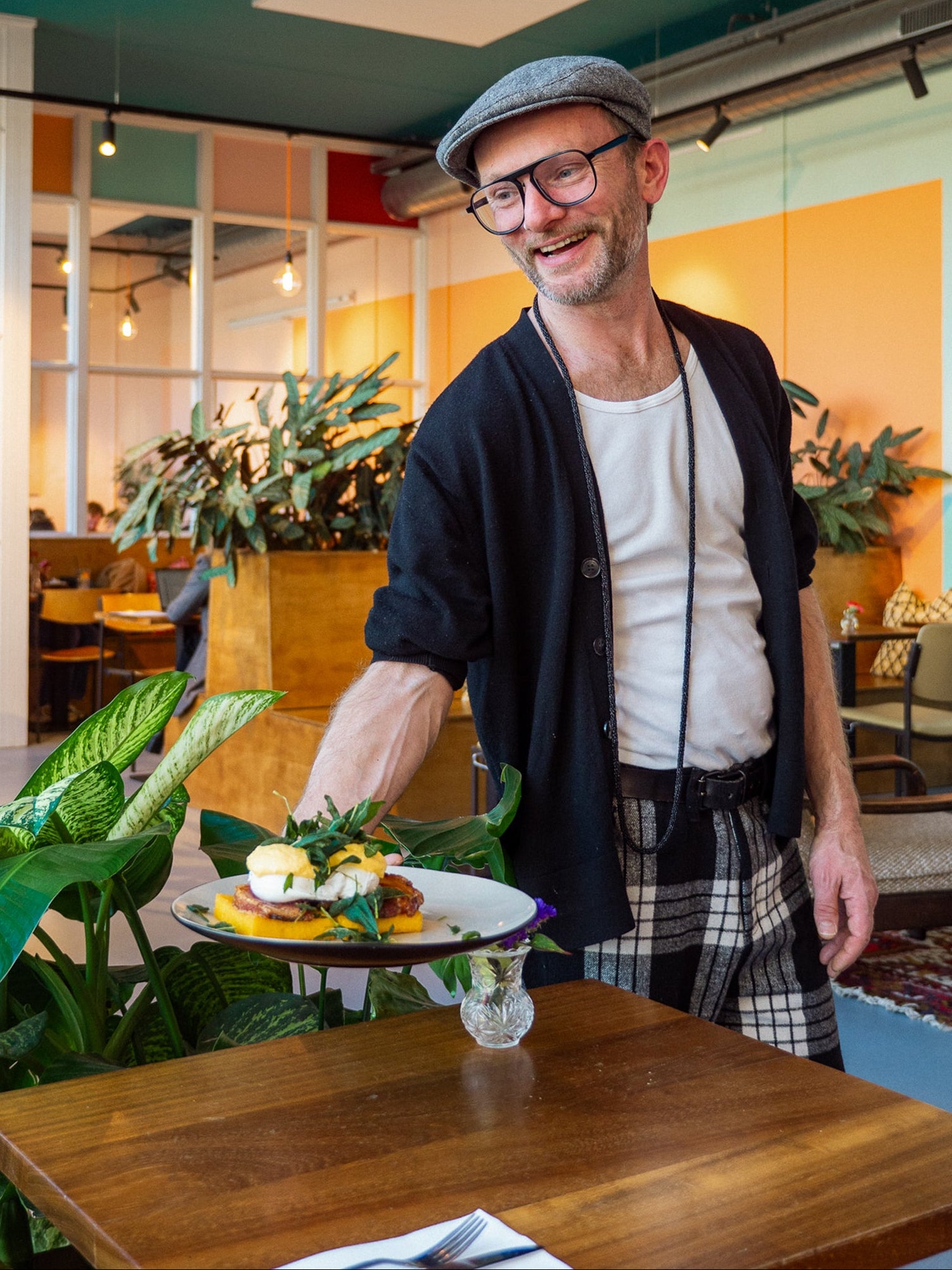 Man in a restaurant holding a plate of food, surrounded by plants and tables.