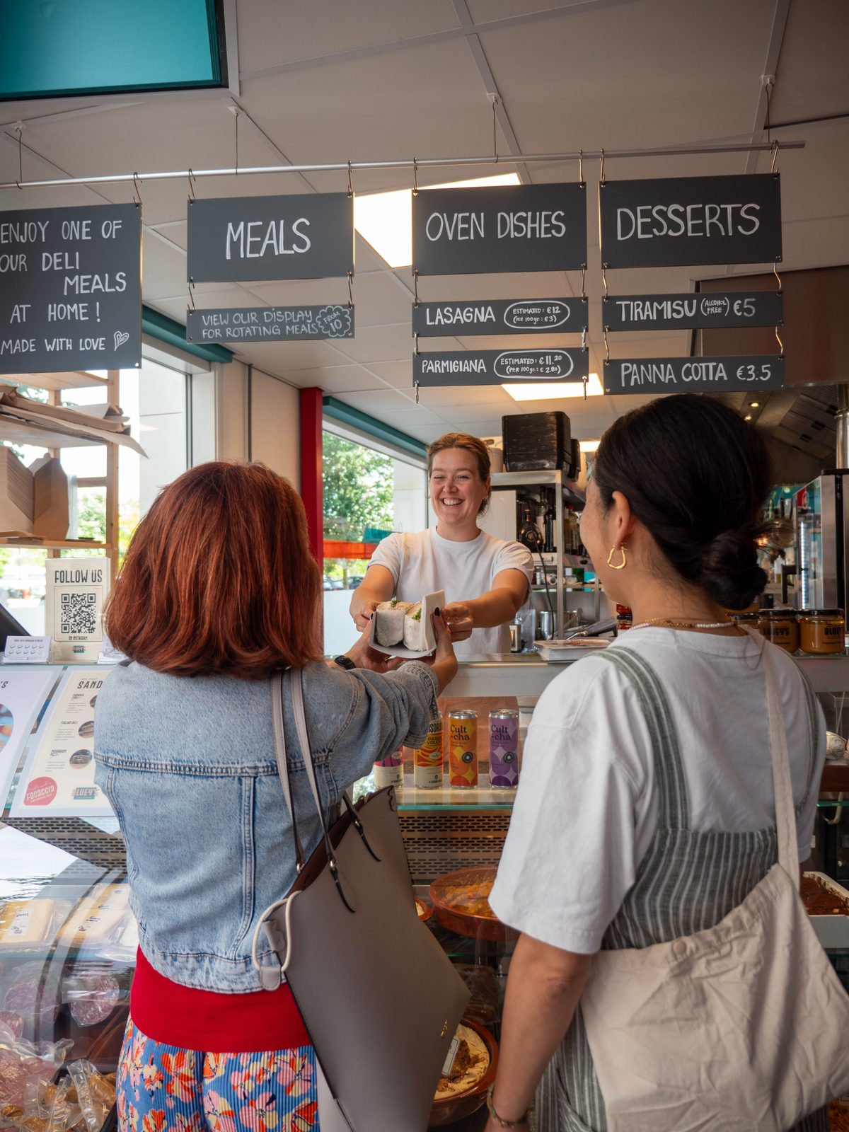 A smiling staff member handing a wrapped takeaway sandwich across the counter to customers at Bluey's deli in Amsterdam