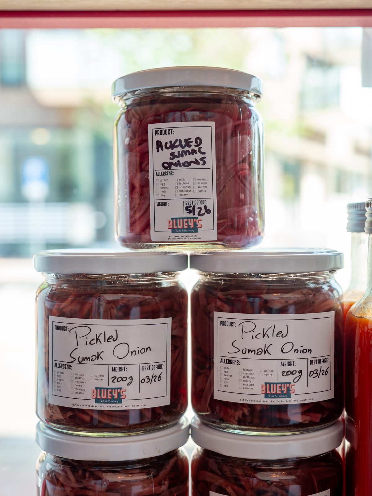 Stacked glass jars of Bluey's homemade pickled sumac onions with handwritten labels, displayed in the window of the deli in Amsterdam