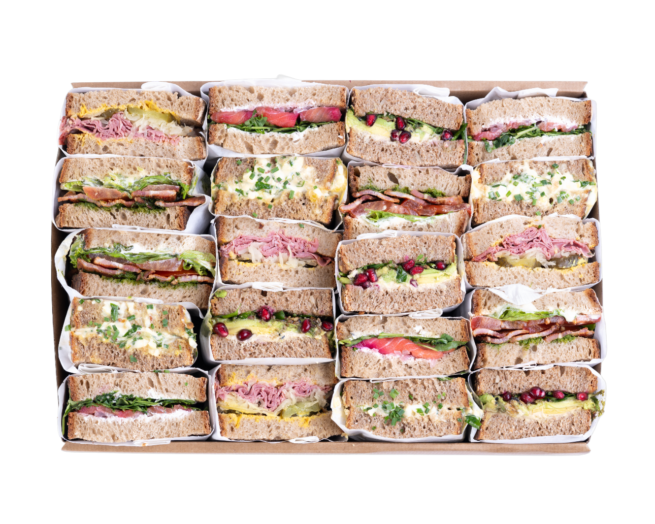 A boxed assortment of catering sandwiches arranged in a grid on a white background. The sandwiches are made with brown whole wheat bread and filled with various ingredients including ham, cheese, greens, and pomegranate seeds.